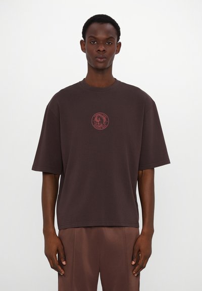 Brun oversized t-skjorte laget av bomull, med en sirkulær rød grafisk logo på brystet, korte ermer og en avslappet passform.