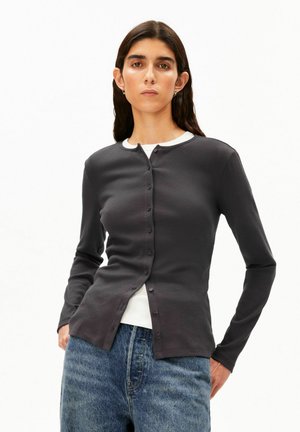 Junge Frau mit dunklen Haaren, die eine dunkelgraue, geknöpfte Strickjacke über einem weißen Top und blauen Jeans trägt, steht mit den Händen in den Taschen.