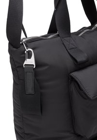 Sort polstret tote taske med lynlåslukning, der har en sølvmetalclip, stofrem og en frontlomme med klap. Teksturerede detaljer og syninger er synlige.