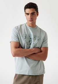 Tricou din bumbac de culoare bleu deschis, cu un grafic triunghiular "GUESS". Mâneci scurte, decolteu rotund și o croială confortabilă.