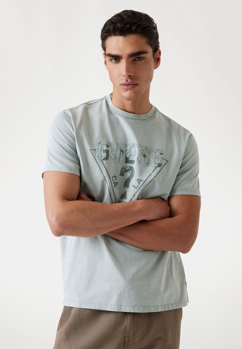 Tricou din bumbac de culoare bleu deschis, cu un grafic triunghiular "GUESS". Mâneci scurte, decolteu rotund și o croială confortabilă.