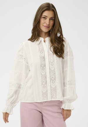 Kobieta o długich brązowych włosach, ubrana w białą bluzkę z koronkowymi zdobieniami i bufiastymi rękawami oraz jasnoróżowe spodnie, delikatnie uśmiechająca się na jednolitym tle.