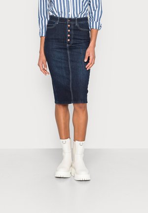 Kvinde iført en mørkeblå knappet denim blyantsnederdel, stribet blå og hvid skjorte og hvide chunky ankelstøvler på en ensfarvet baggrund.