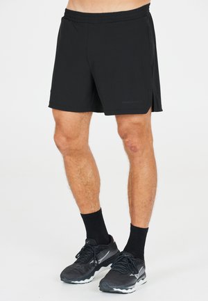 Zwarte sportshorts van lichtgewicht materiaal met een elastische tailleband en zijsplitten voor bewegingsvrijheid. Gecombineerd met zwarte schoenen en sokken.