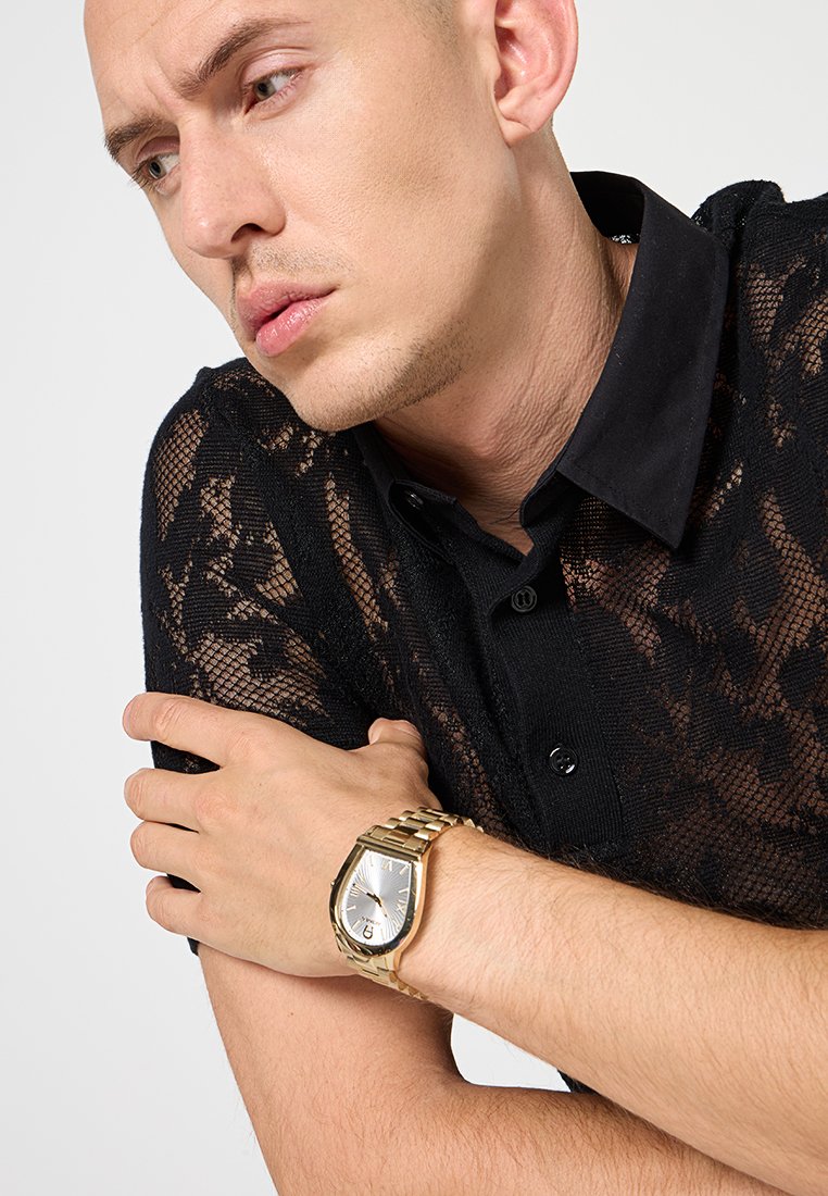 Schwarzes Spitzenhemd mit Kragen, das eine durchsichtige Textur aufweist. Goldene Uhr am Handgelenk mit quadratischem Ziffernblatt und Gliederarmband. Die Arme vor der Brust verschränkt.