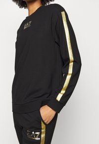 Svart sweatshirt med långa ärmar som har guldglittrande ränder och EA7-logotyp. Mjuk tygkvalitet med en avslappnad passform.