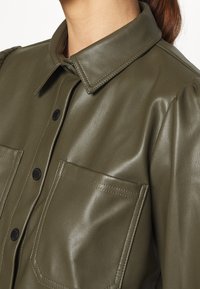Giacca camicia in finta pelle verde oliva, con colletto a punta, bottoni a scatto frontali e due tasche sul petto. Texture liscia con spalle imbottite.
