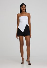 Top bianco senza spalline con orlo asimmetrico abbinato a pantaloni corti neri. Il modello indossa tacchi neri con cinturini. Design semplice e moderno con colori a contrasto.