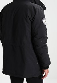 Homme portant une veste d'hiver noire avec un patch circulaire de marque sur le haut de la manche gauche, debout devant un fond uni.