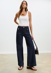 Jeans in denim scuro a gamba larga con accessori dorati, tasche frontali e cuciture a contrasto. Abbinati a un top bianco senza maniche e sandali marroni.