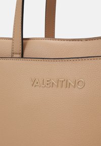 Krémová kožená taška přes rameno s texturovaným povrchem, hladkými popruhy a embosovaným logem "VALENTINO". Černé stehy obrysují horní hranu.