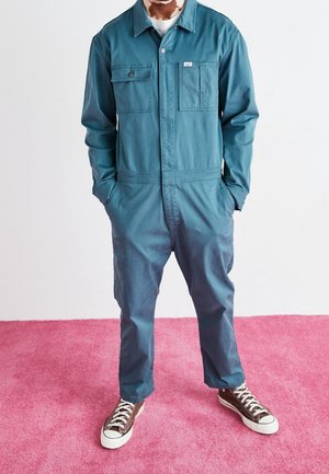 Ein Mann trägt einen blauen Overall mit Brusttaschen und braune High-Top-Sneaker, steht auf einem pinkfarbenen Teppich vor einer weißen Wand.
