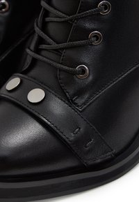Gros plan sur une botte en cuir noire avec de gros lacets, des œillets métalliques et deux clous ronds en argent sur une sangle en cuir à l'avant.