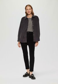 Chemise-veston en velours côtelé gris oversize, pantalon noir ajusté et mocassins noirs avec détail en chaîne. Texture lisse, fermetures à boutons sur la veste.