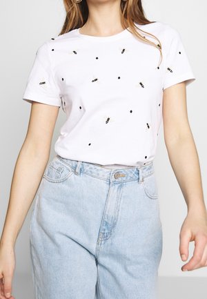 Femme portant un t-shirt blanc à manches courtes avec de petits imprimés d’abeilles et de points, rentrée dans un jean taille haute bleu clair.