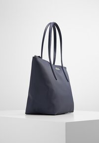 Bolsa de mano azul marino hecha de material sintético texturizado, con dos correas largas y cierre de cremallera. Presenta un pequeño logo verde en la parte frontal.