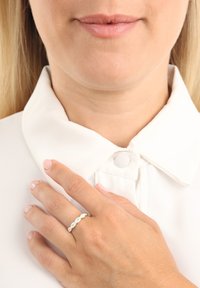 Vit skjorta med knäppbar krage; hand med en ring av guld och silver i växlande mönster med en slät, vågig design och utan synliga ädelstenar.