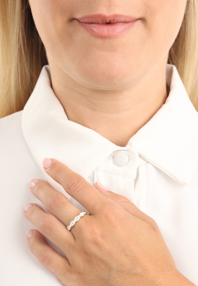 Vit skjorta med knäppbar krage; hand med en ring av guld och silver i växlande mönster med en slät, vågig design och utan synliga ädelstenar.