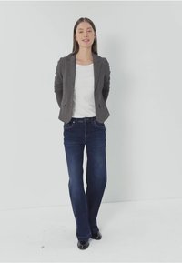 Grauer Blazer über einem weißen Oberteil, kombiniert mit dunkelblauen Schlagjeans. Das Outfit weist klassische Denim-Details und schwarze Schuhe auf.