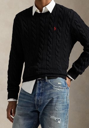 Persoon draagt een zwarte kabelgebreide trui over een wit overhemd, gecombineerd met versleten blauwe spijkerbroek, hand rustend in de zak.