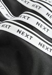 Crna tkanina s bijelim elastičnim pojasom. Pojas sadrži ponovljeni "NEXT" brend u crnom slovima na glatkoj, teksturiranoj pozadini.