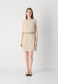 Beige tweed-mekko, jossa on suora siluetti, hihaton muotoilu ja vaalea vyö. Yhdistetty mustiin terävään kärkeen oleviin korkokenkiin.