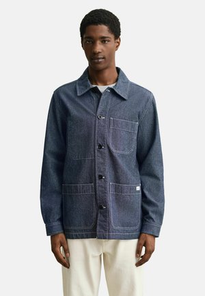 Homme portant une veste en denim bleu foncé boutonnée avec trois poches avant et un pantalon de couleur crème, debout devant un fond clair uni.