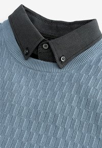 Pull en maille bleu clair avec un col en chevrons noir, présentant des accents de boutons et un col côtelé.