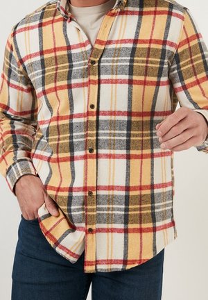 Mężczyzna w żółto-czerwono-czarno-białej koszuli w kratę zapinanej na guziki, na beżowej koszulce, z jedną ręką w kieszeni, a drugą uniesioną.