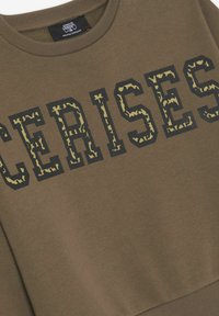 Sweatshirt vert olive avec un col et des poignets côtelés. Présente du texte noir en gras "CERISES" avec un motif camouflage jaune. Tissu doux.