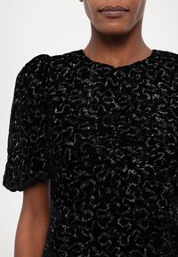 Femme portant une robe en velours texturé noire avec des manches courtes bouffantes et un motif léopard subtil.