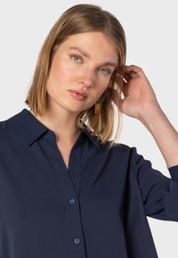 Camicia button-up blu navy con colletto, realizzata in tessuto morbido, maniche lunghe e una leggera lucentezza. Orecchini quadrati dorati aggiungono contrasto.
