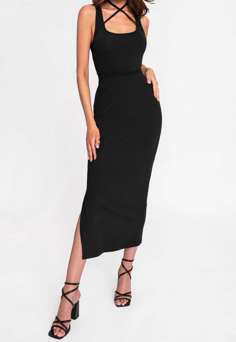 Femme portant une robe noire ajustée sans manches avec des bretelles croisées et des sandales à talons hauts noires à lanières, posant devant un fond blanc.