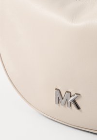Lys beige læderhåndtaske med en glat overflade og rund form; har et metallisk "MK"-logo på forsiden.