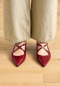 Red patent leather pointed-toe shoes with cross strap detail, featuring a smooth texture and a low heel. Worn with wide beige pants.