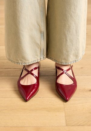 Chaussures à bouts pointus en cuir verni rouge avec brides croisées et côtés ajourés, portées avec un pantalon large en jean clair sur un sol en bois.