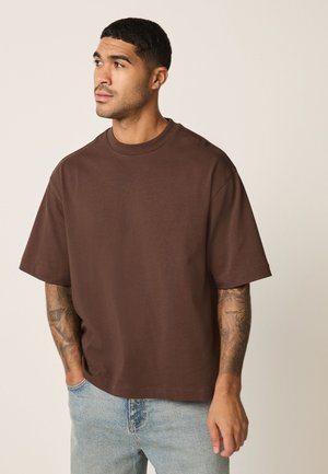 Bărbat cu tatuaje, purtând un tricou maro supradimensionat și blugi bleu deschis, uitându-se în lateral cu o mână în buzunar.