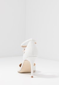 Chaussure à talons hauts blanche avec une surface texturée, une sangle à la cheville et un talon élancé. La semelle extérieure est de couleur nude avec un léger accent sur le talon.