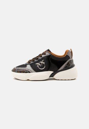 Zapatilla negra con detalles en marrón y gris, suela blanca gruesa, paneles de malla y cuero, y un logo decorativo plateado en el lateral.