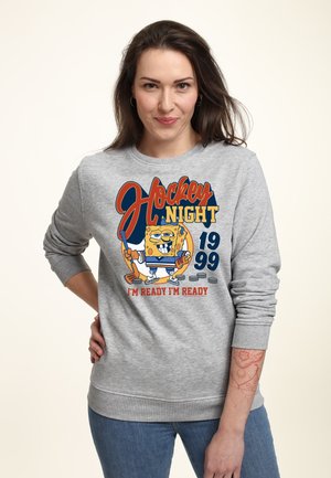 Femme aux cheveux bruns portant un sweat-shirt gris avec un personnage de dessin animé en équipement de hockey, texte « Hockey Night 1999 I'm Ready I'm Ready »