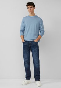 Hellblauer Strickpullover mit horizontalen Streifen, kombiniert mit dunkelblauen Jeans und weißen Turnschuhen. Hände in den Taschen, stehende Pose.