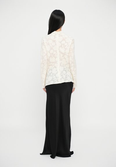 Blusa bianca trasparente con motivi floreali abbinata a una lunga gonna nera liscia. La blusa ha maniche lunghe e un taglio dritto.