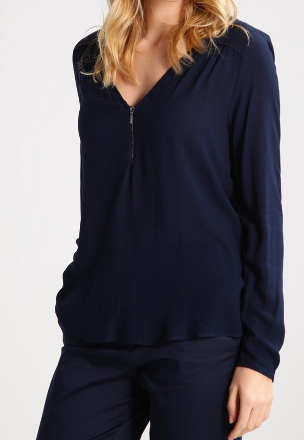 Femme portant un chemisier bleu marine à manches longues avec une fermeture éclair à l'avant et un pantalon bleu marine assorti sur fond blanc.