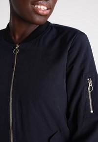 Marinblå bomberjacka med ribbad krage, guldrivknapp och sidofickor. Har en ficka med dragkedja på ärmen för extra förvaring.