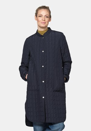 Woman wearing a long navy quilted coat with snap buttons, standing with hands in pockets against a plain light background.