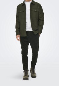 Olijfgroen button-up jas over een wit T-shirt, gecombineerd met donkergroene corduroy broek en veterschoenen, wat een gelaagde, gestructureerde look laat zien.
