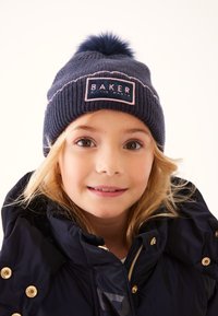 Laivastonsininen neulottu pipo, jossa on päällä pom-pom ja suorakulmainen merkki, jossa lukee "BAKER" vaaleanpunaisella. Pehmeä koostumus ja resorikuvio.