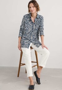 Navy-blaues, mit Blumenmuster versehenes, langärmliges Hemd mit Knöpfen, kombiniert mit weißen hochgekrempelten Hosen und schwarzen Slippern.
