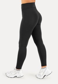 Frau trägt schwarze Leggings mit hoher Taille und weiße Sportschuhe und steht auf einem schlichten hellen Hintergrund.
