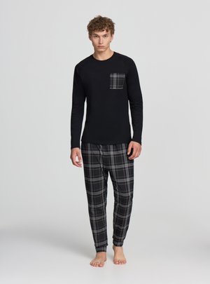 Młody mężczyzna stojący boso, ubrany w czarną bluzę z długim rękawem z kieszenią na piersi w kratę oraz dopasowane czarno-szare spodnie w kratę.